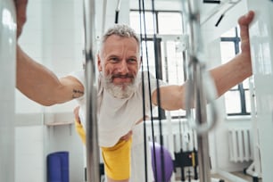 Smiling pleased gray-haired bearded man doing a strength training exercise assisted by a physical therapist
