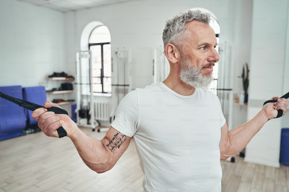 Retrato de cintura para cima de um esportista de cabelos grisalhos e calmo fazendo os cachos laterais do cabo do bíceps em pé
