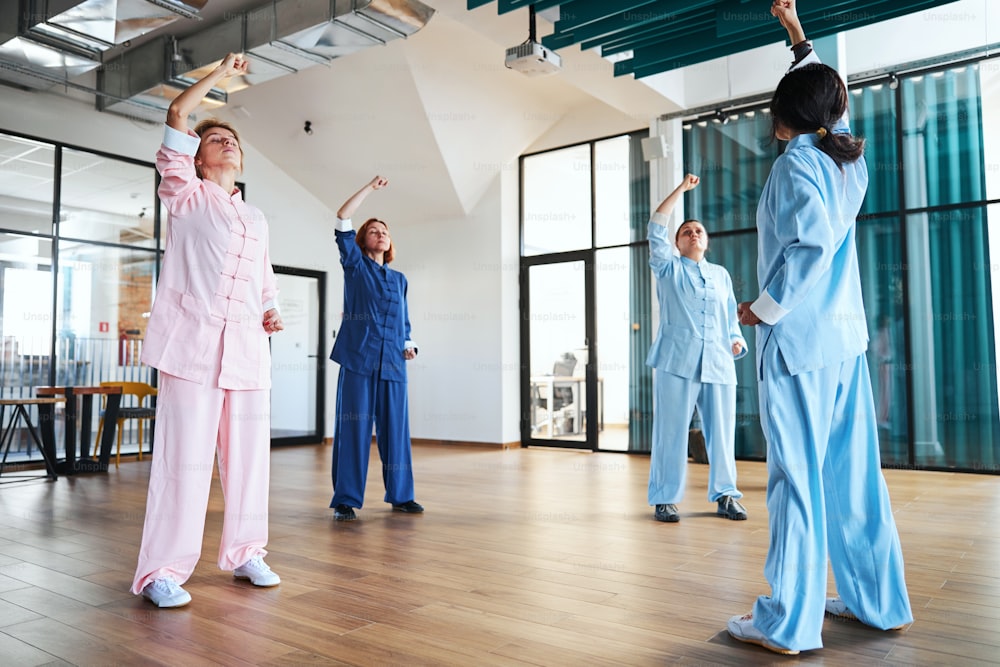 Group of adult women practicing qigong in a spacious room and wearing Chinese clothes