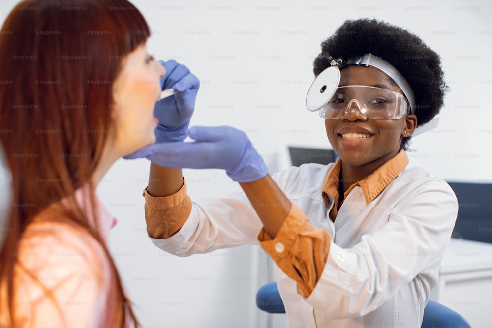 Positive grinning young African American lady doctor otorhinolaryngologist, wearing uniform, frontal reflector and eyglasses, checking throat of her female patient, using tongue depressor