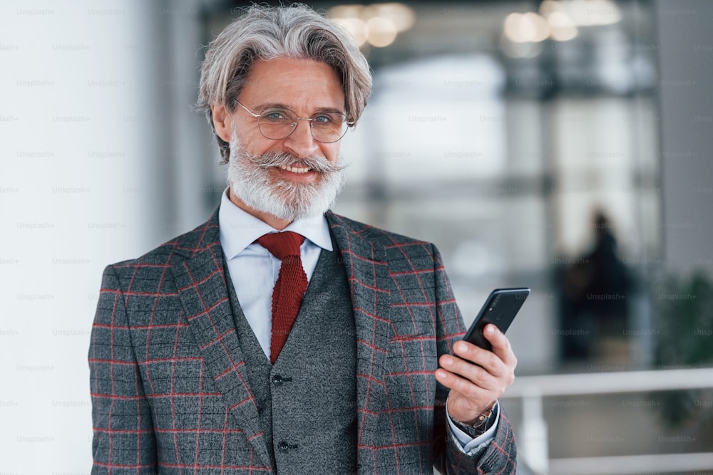Senior businessman in suit and tie with gray hair and beard standing indoors with phone.