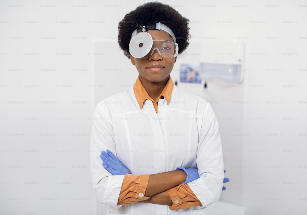 Portrait of young pleasant African American woman, confident ENT otolaryngologist doctor, wearing white lab coat, head frontal reflector and protective glasses, posing to camera with arms crossed