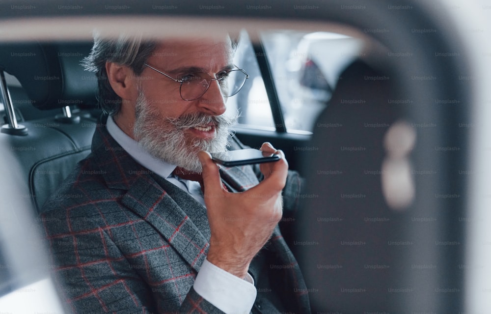 Modern stylish senior man with grey hair and mustache talking by the phone inside automobile.