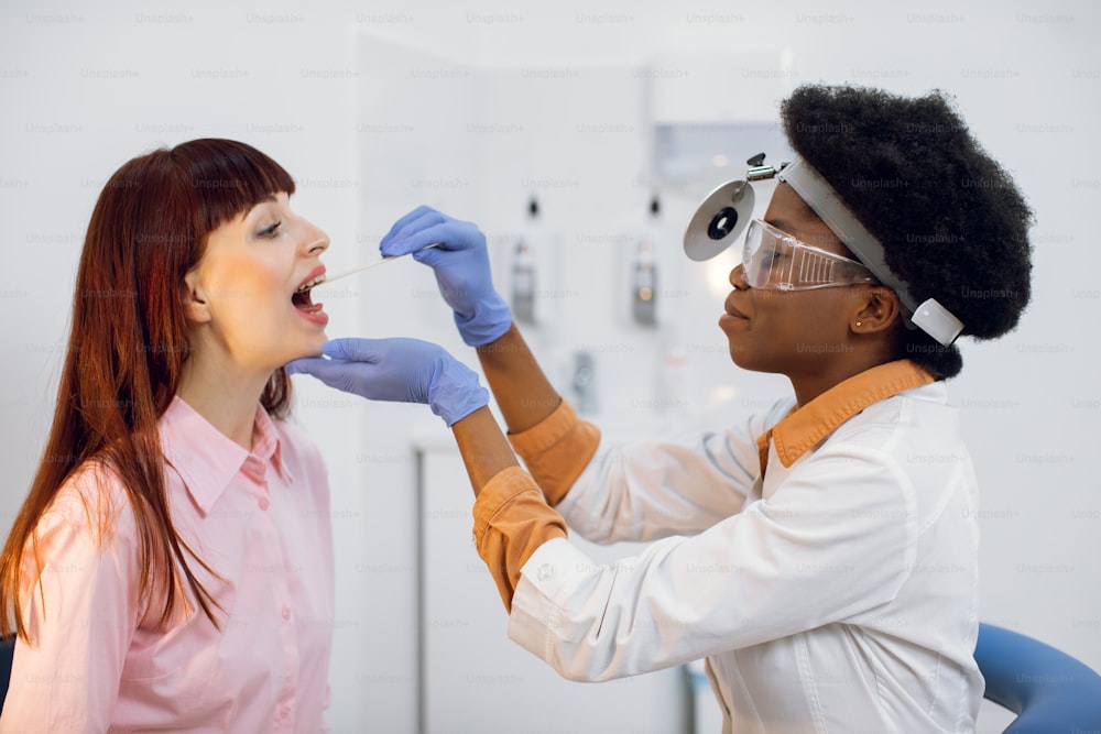 Joven médico otorrinolaringólogo afroamericano examinando la laringe de una joven mujer europea en un consultorio moderno. Médico de cabecera terapeuta de mujer negra, usando un depresor de madera, comprobando el dolor de garganta de una paciente joven