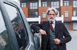 Modischer älterer Mann mit grauen Haaren und Bart, der sich an sein Auto lehnt und Schlüssel hält.