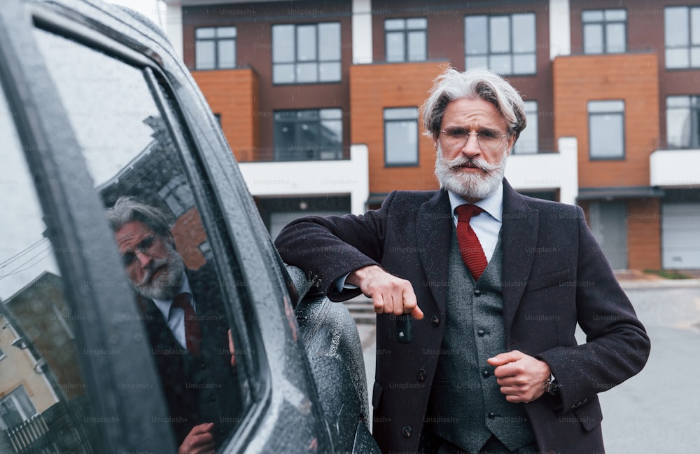 Fashionable senior man with gray hair and beard leaning on his car and holding keys.