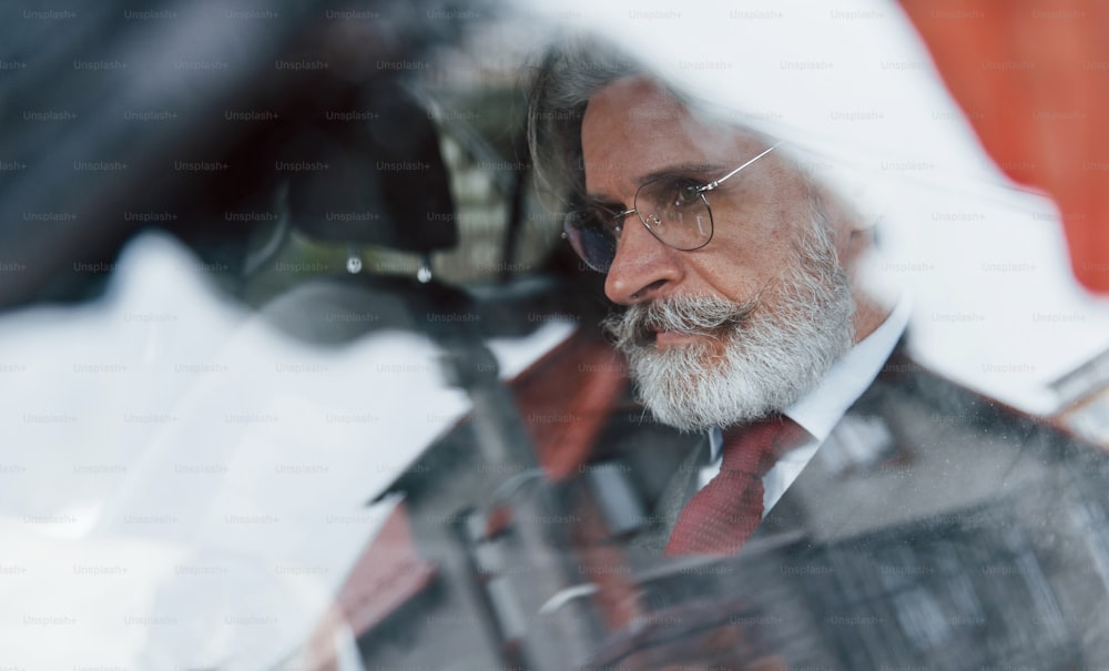 View through the glass of stylish senior man with grey hair and mustache that is in the modern car.