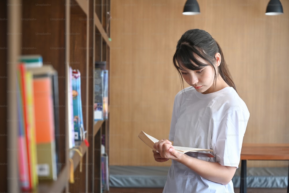 Young female student standing near bookshelf in library and reading a book.