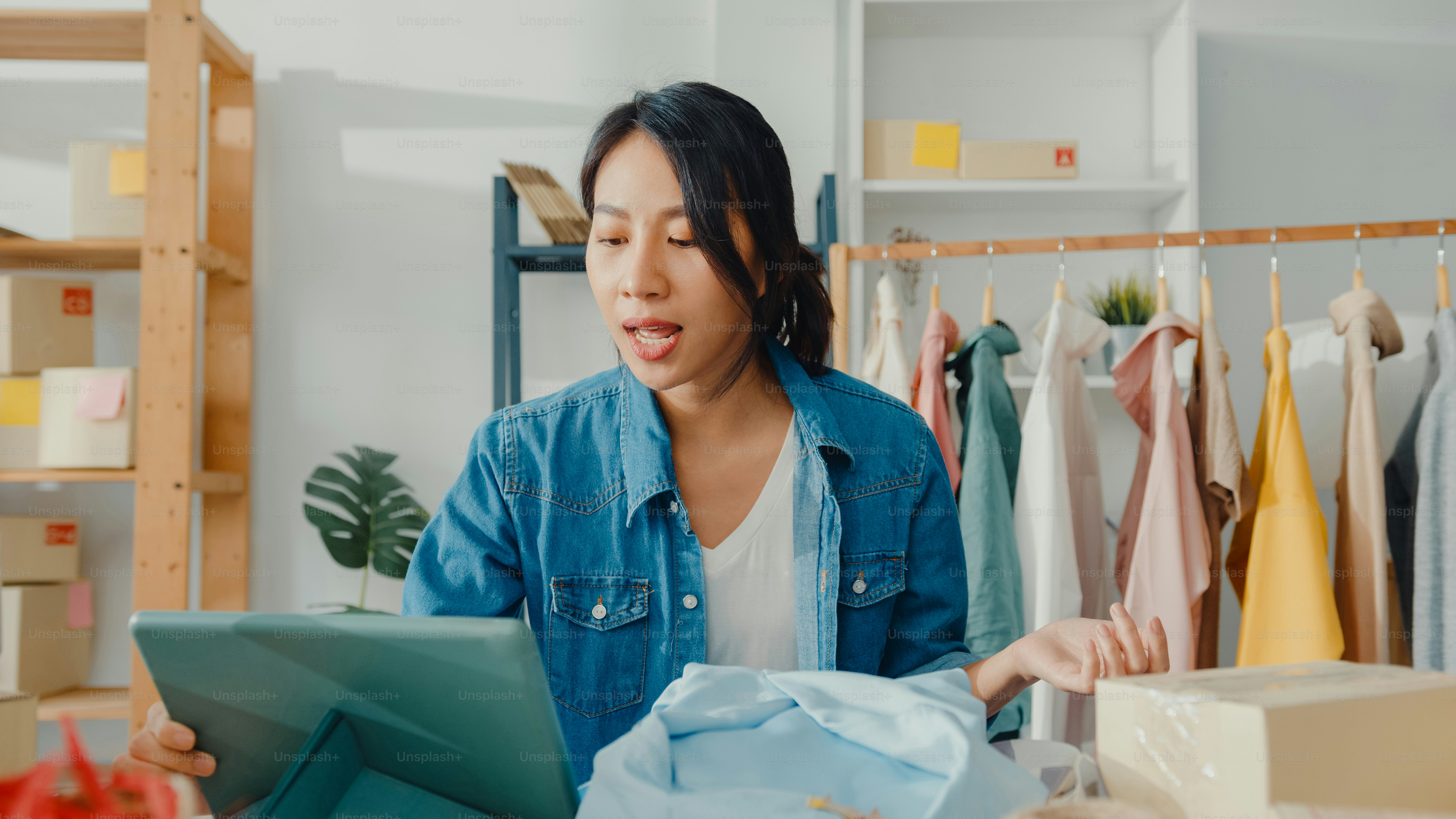 Young Asia lady fashion designer using mobile phone receiving purchase order and showing clothes recording video live streaming online at shop. Small business owner, online market delivery concept.