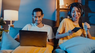 Freelance asia couple man and woman in casual hasband work laptop computer at night enjoy happy relax with wife wear headphone listen music in smartphone sing dance on couch at house, Married life.