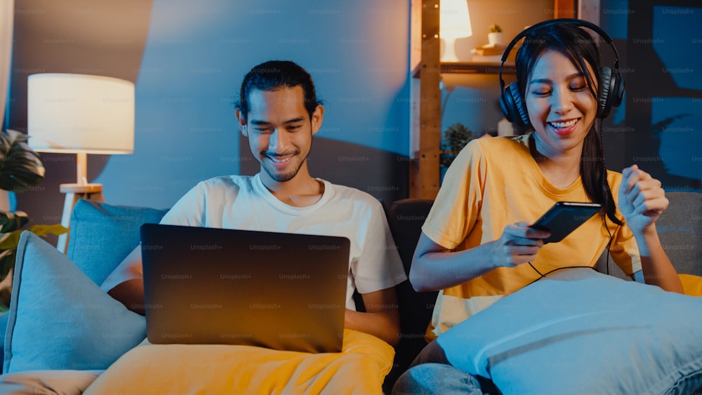 Freelance asia couple man and woman in casual hasband work laptop computer at night enjoy happy relax with wife wear headphone listen music in smartphone sing dance on couch at house, Married life.