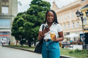 Cheerful african american woman using smartphone and drinking coffee takeaway while walking in the city on a sunny day