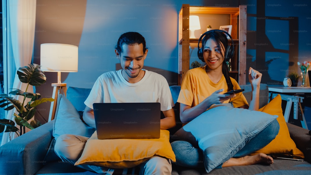 Freelance asia couple man and woman in casual hasband work laptop computer at night enjoy happy relax with wife wear headphone listen music in smartphone sing dance on couch at house, Married life.