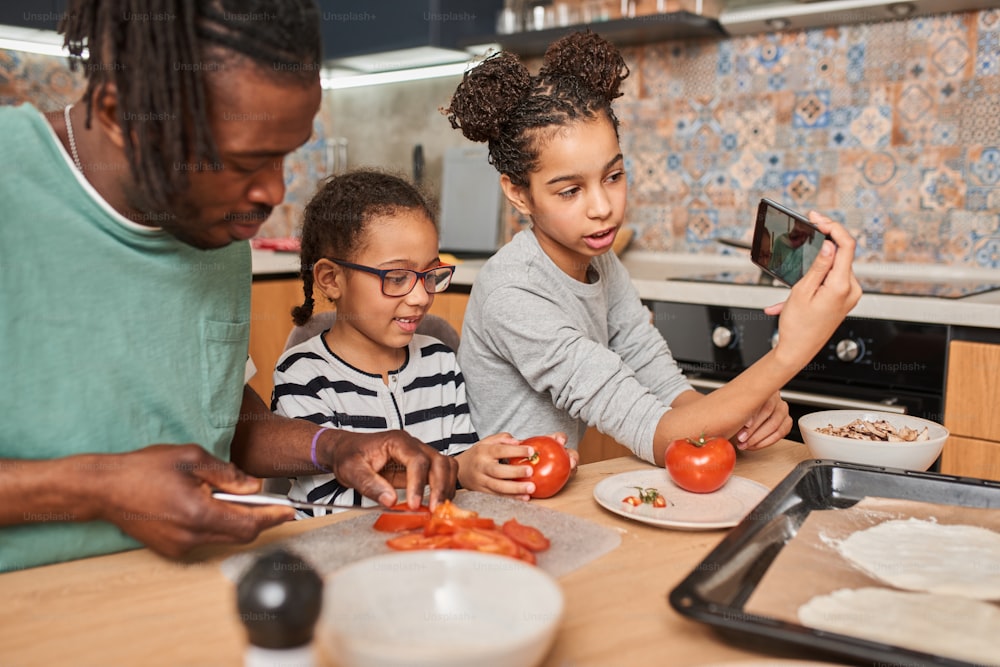 スマートフォンを持ち、自宅のキッチンでピザを準備しながら妹と一緒にトマトを刻む幸せな父親と自撮りをしている笑顔の多民族の女の子の腰のアップポートレートビュー