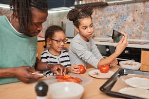 Waist up portrait view of the smiling multiracial girl holding smartphone and making selfie with her happy father who chopping tomato with her little sister while preparing pizza in kitchen at home