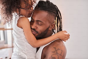 Childhood. Cute small girl feeling satisfied and happy while spending day with her father. Strong tattooed father embracing his lovely daughter with closed eyes