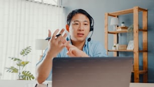 Young Asia businessman wear headphones using laptop talk to colleagues about plan in video call while work from home at living room. Self-isolation, social distancing, quarantine for covid prevention.