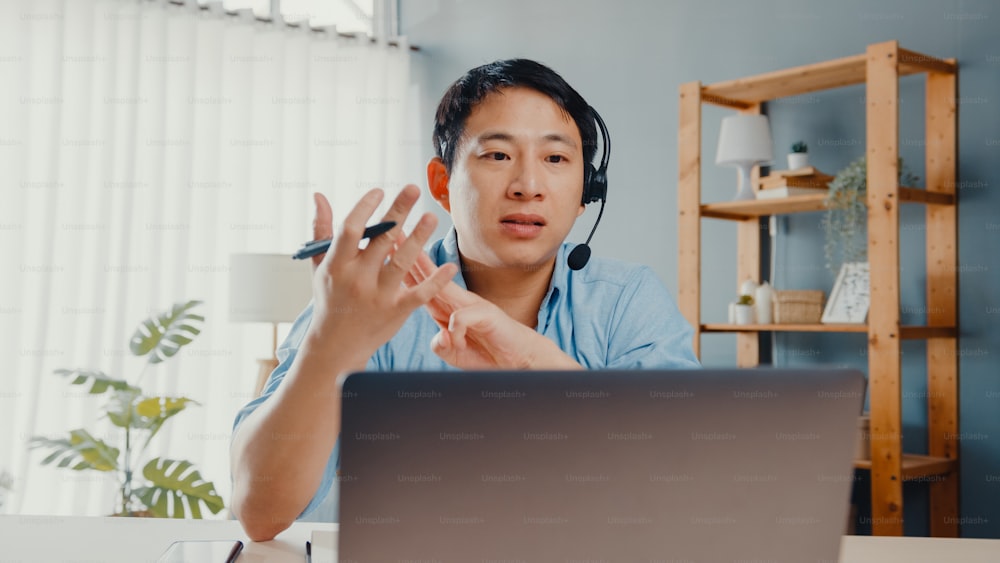 Il giovane uomo d'affari asiatico indossa le cuffie usando il computer portatile parla con i colleghi del piano in videochiamata mentre lavora da casa in salotto. Autoisolamento, distanziamento sociale, quarantena per la prevenzione del covid.