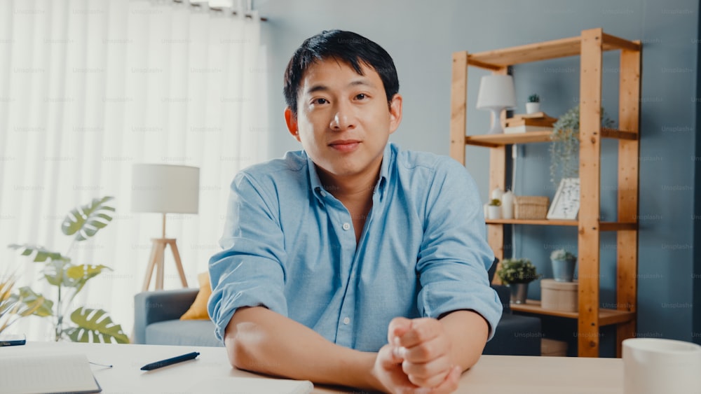 Ein junger asiatischer Geschäftsmann mit Computer-Laptop spricht mit Kollegen über den Plan in einem Videoanruf-Meeting, während er von zu Hause aus im Wohnzimmer arbeitet. Selbstisolation, soziale Distanzierung, Quarantäne für Coronavirus.