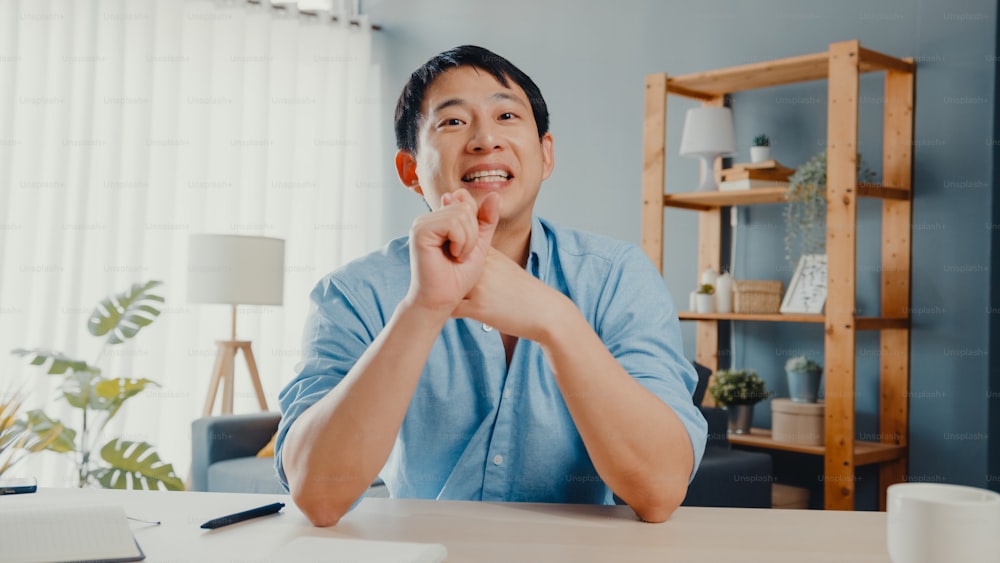 Ein junger asiatischer Geschäftsmann mit Computer-Laptop spricht mit Kollegen über den Plan in einem Videoanruf-Meeting, während er von zu Hause aus im Wohnzimmer arbeitet. Selbstisolation, soziale Distanzierung, Quarantäne für Coronavirus.