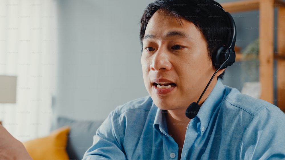 Junge asiatische Geschäftsleute tragen Kopfhörer mit Laptop Sprechen Sie mit Kollegen über Plan in Videoanrufen, während Sie von zu Hause aus im Wohnzimmer arbeiten. Selbstisolation, soziale Distanzierung, Quarantäne zur Covid-Prävention.