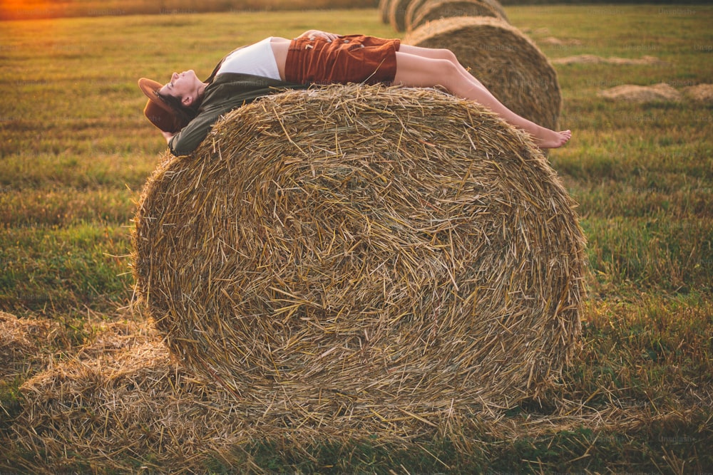 夏の野原で夕暮れの光の中で干し草の山に横たわっている帽子をかぶった美しい屈託のない女性。田舎の干し草の俵でリラックスしている若い幸せな女性。雰囲気のある静かなひととき