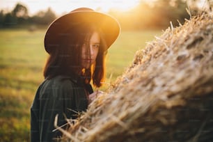 Beautiful stylish woman in hat posing at haystacks in sunset light in summer field. Atmospheric tranquil moment in countryside. Young female relaxing at hay bale in evening warm sunshine