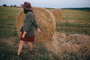 Beautiful stylish woman in hat dancing at hay bales in summer evening field. Carefree happy moment, vacation in countryside. Happy young female relaxing and having fun at haystacks