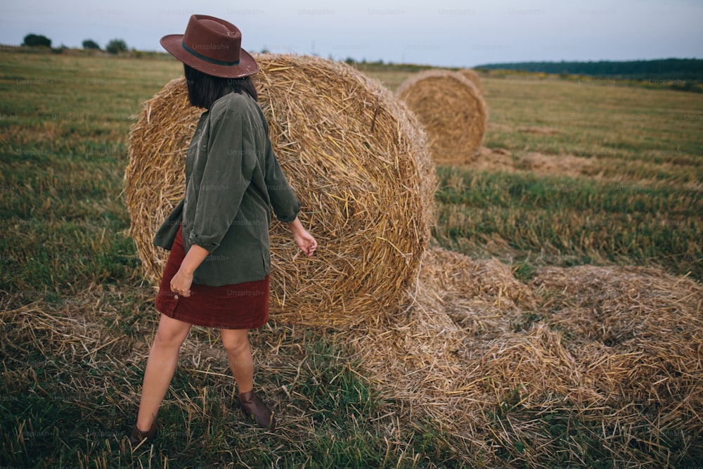 Beautiful stylish woman in hat dancing at hay bales in summer evening field. Carefree happy moment, vacation in countryside. Happy young female relaxing and having fun at haystacks