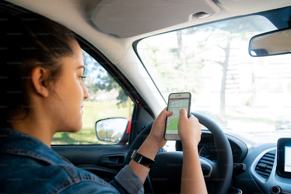 그녀의 차를 운전하는 동안 그녀의 휴대 전화에 GPS 네비게이션 시스템을 사용하는 젊은 여자의 초상화. 운송 및 탐색 개념입니다.