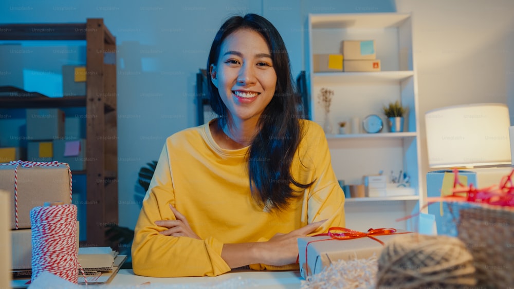 Porträt einer jungen asiatischen Geschäftsfrau, die glücklich lächelt, die Arme verschränkt und in die Kamera schaut, während sie nachts im Home-Office arbeitet. Kleinunternehmer, Online-Markt-Lieferkonzept.