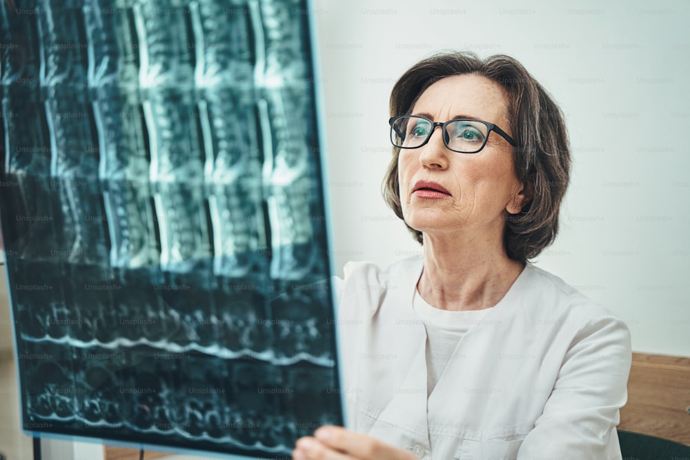 Concentrated experienced mature woman doctor in eyeglasses examining the MRI scans of the lumbar spine