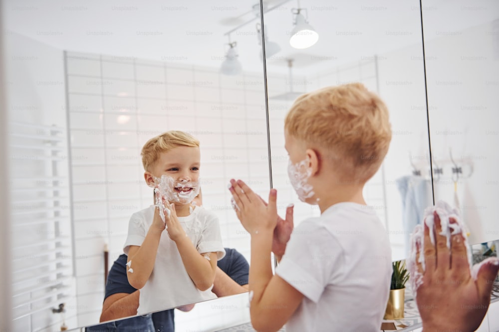 Father with his son is in the bathroom have fun by using shaving gel and looking in the mirror.