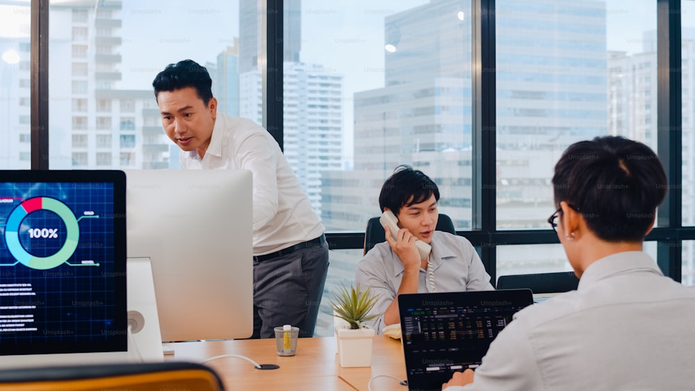 Millennial group of young Asia businesspeople in small modern office. Japanese male boss supervisor teaching intern or new employee Chinese young guy helping with difficult assignment at meeting room.