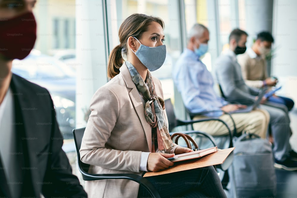 Junge Geschäftsfrau mit Gesichtsschutzmaske, während sie auf ein Vorstellungsgespräch wartet und im Wartezimmer eines Bürogebäudes sitzt.