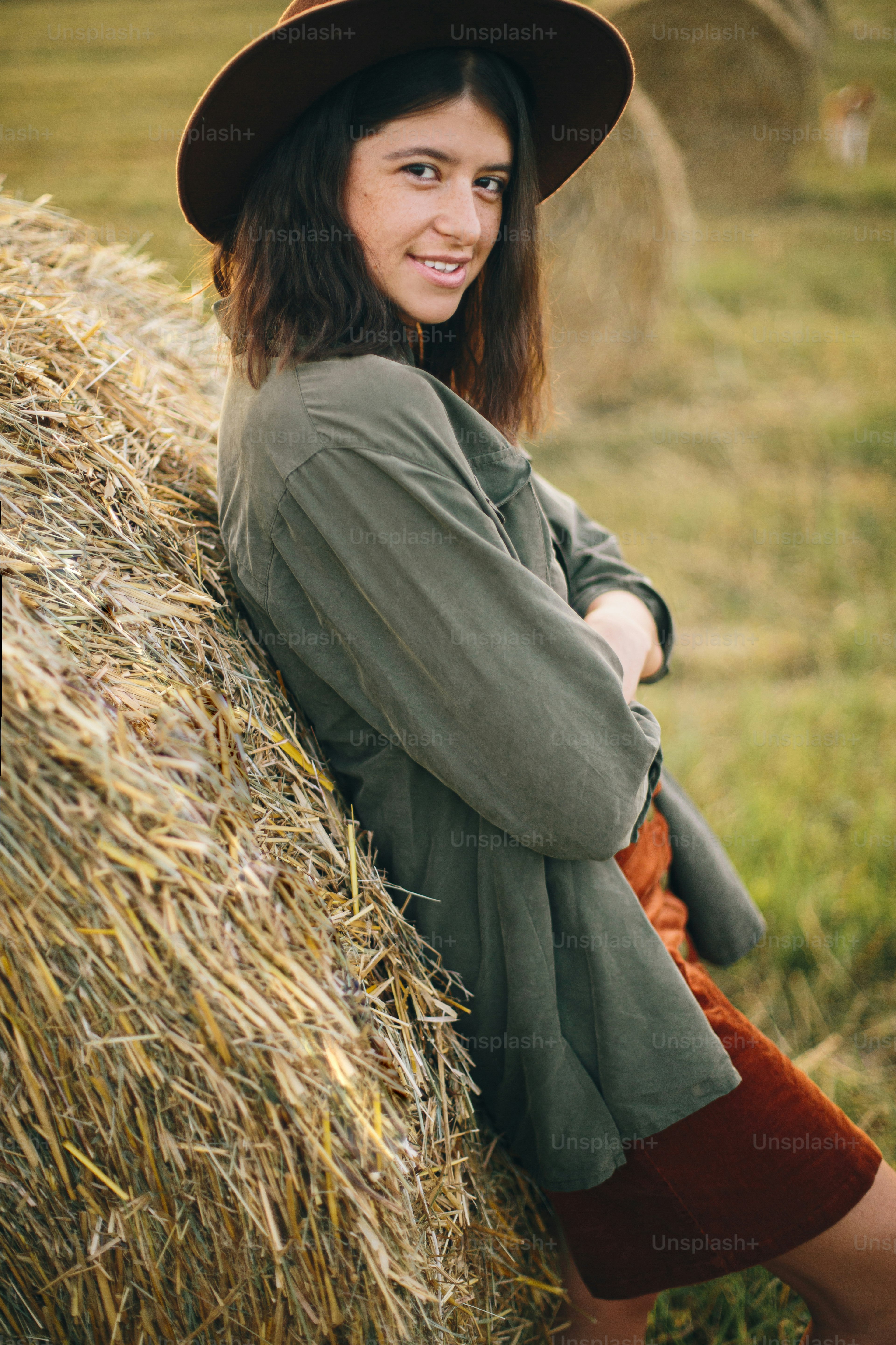 Beautiful stylish woman in hat standing at hay bale in summer evening field. Portrait of young fashionable female relaxing at haystacks, summer vacation in countryside. Tranquility