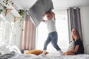 Mother playing pillow fight with her son in bedroom at daytime.