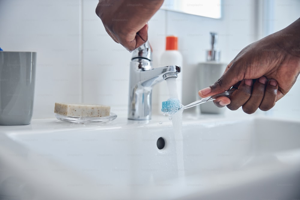 Focused photo on dark-skinned male person that standing near sink and putting brush under the water before cleaning teeth
