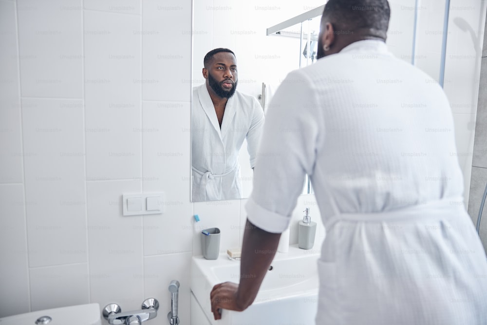 Attentive bearded male person leaning on the sink while preparing for working day in the bathroom