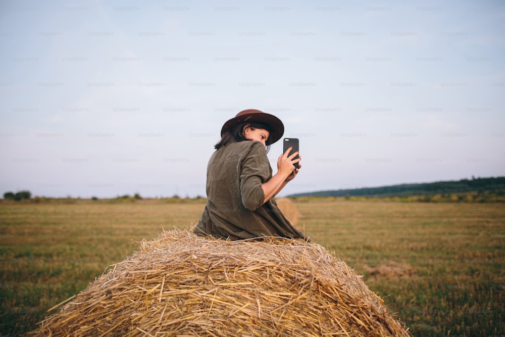 Beautiful stylish woman sitting on haystack and taking photo of sunset on phone in summer field. Young female holding smartphone and capturing evening in warm sunshine. Atmospheric moment