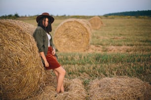 Stylish woman in hat standing at hay bale in summer evening in field. Atmospheric tranquil moment. Young fashionable female relaxing at haystack, summer vacation in countryside. Rural slow life