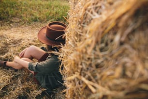 Beautiful stylish woman in hat sitting at haystacks in evening summer field. Atmospheric tranquil moment in countryside. Young fashionable female relaxing at hay bale, top view