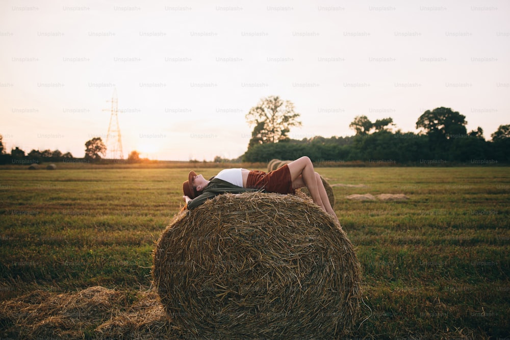 Bella donna spensierata in cappello sdraiata sul pagliaio nella luce del tramonto che si gode la sera nel campo estivo. Giovane femmina felice che si rilassa sulla balla di fieno in campagna. Momento di tranquillità atmosferica