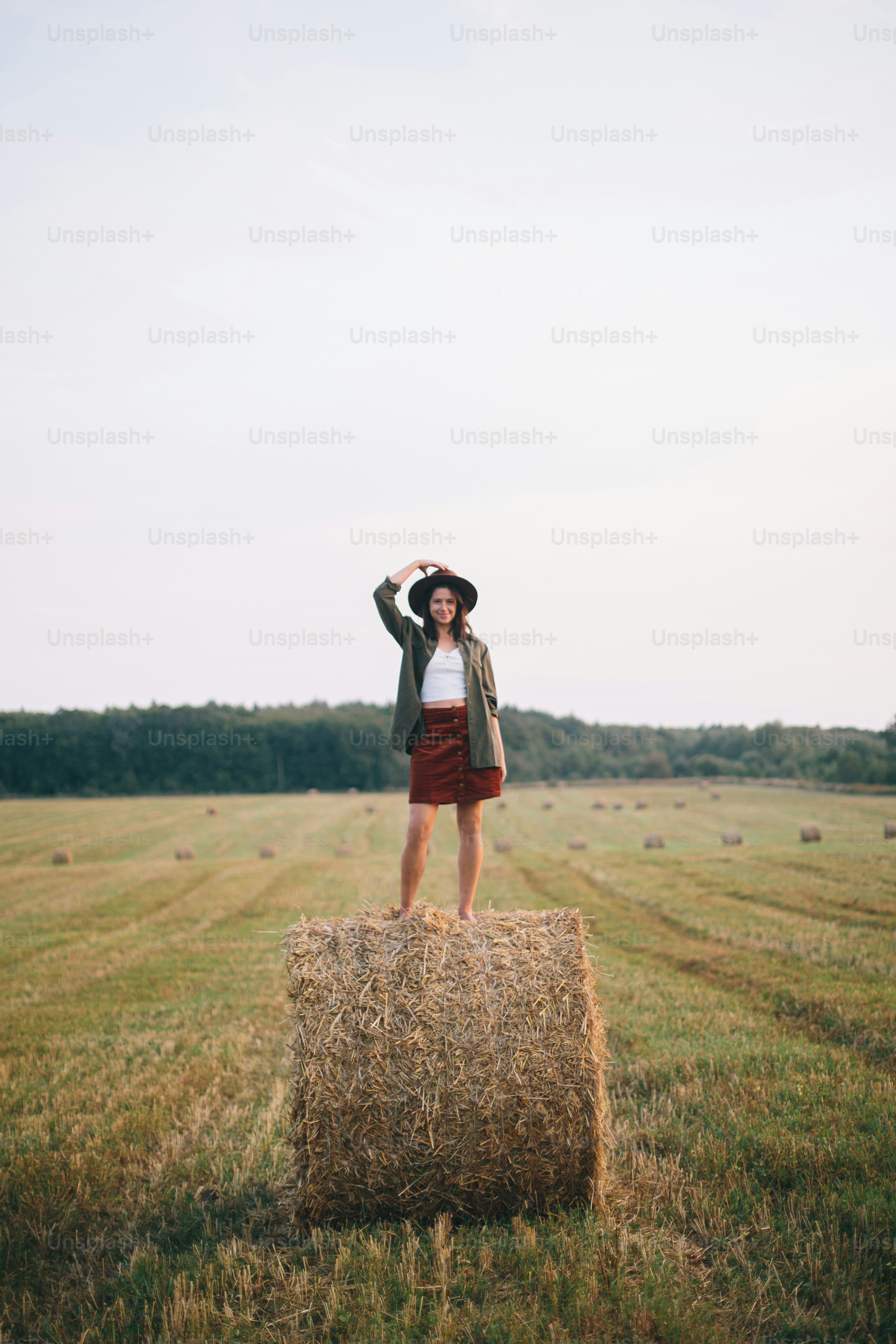 Bella donna spensierata in cappello in piedi sul pagliaio che si gode la sera nel campo estivo. Giovane femmina felice che si rilassa sulla balla di fieno in campagna. Momento di tranquillità atmosferica