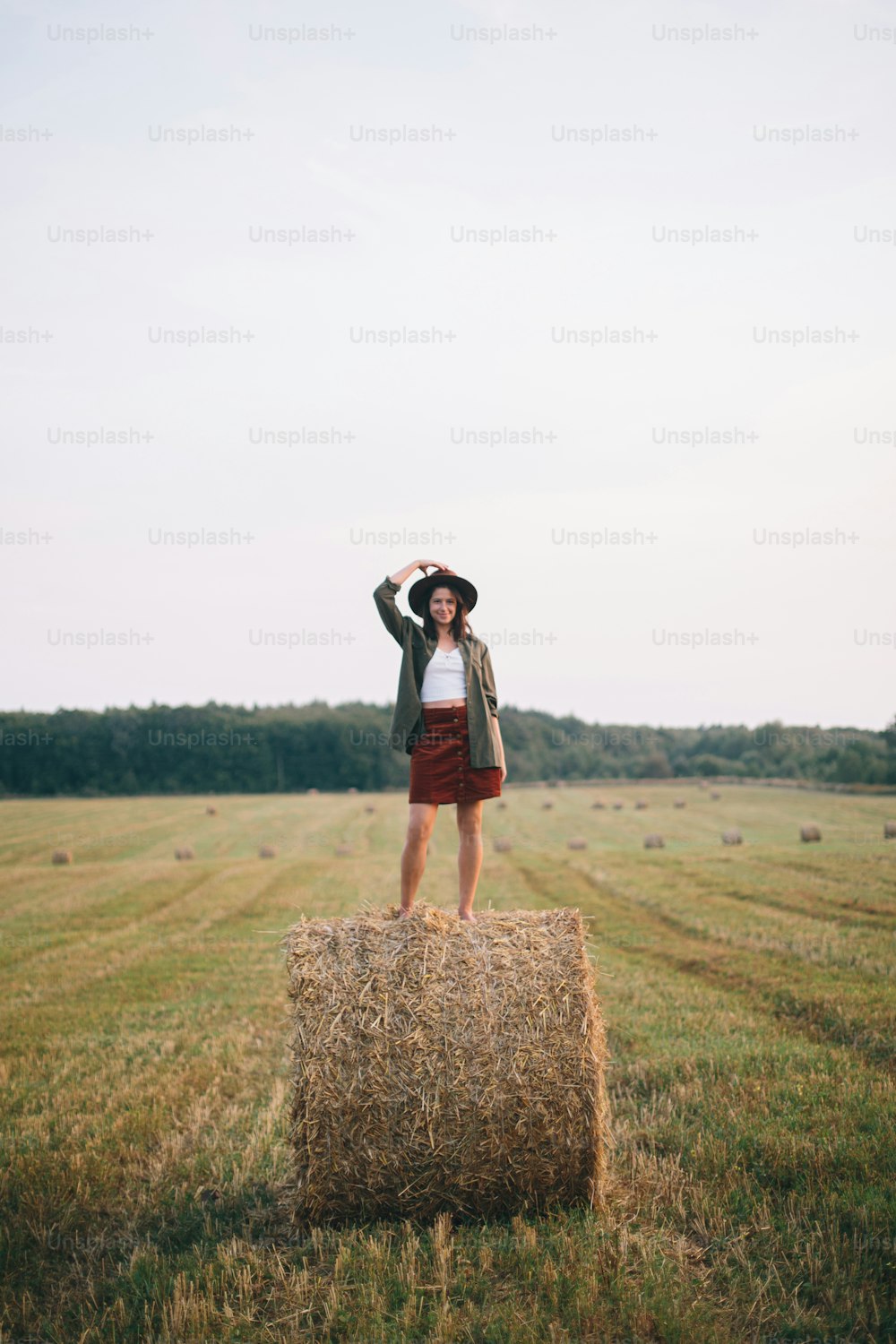 Belle femme insouciante en chapeau debout sur une botte de foin profitant d’une soirée dans un champ d’été. Jeune femme heureuse se relaxant sur une balle de foin à la campagne. Moment de tranquillité atmosphérique