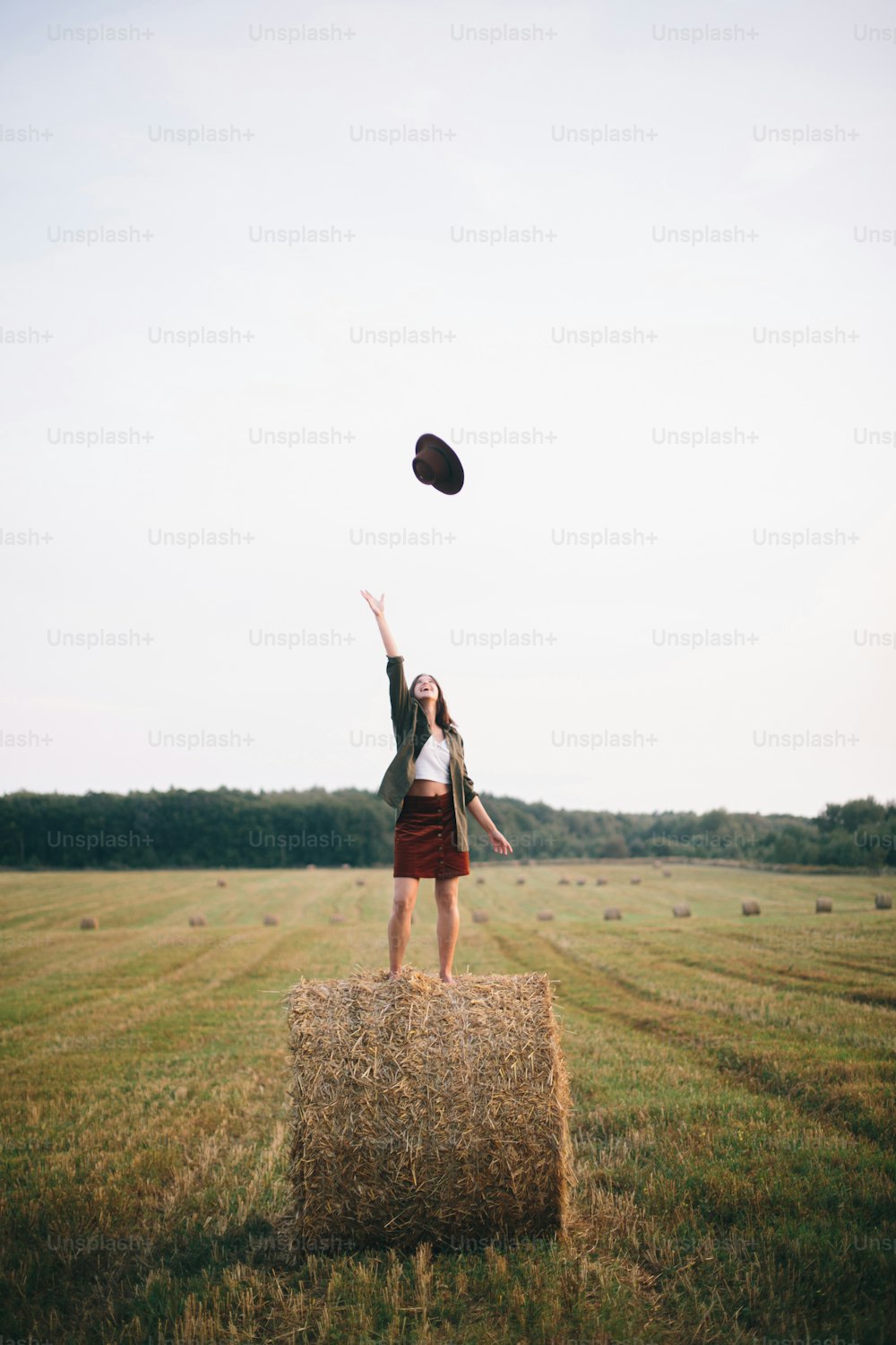 Beautiful carefree woman throw up hat in the sky while standing on haystack in evening summer field. Happiness. Young happy female  having fun on hay bales in countryside. Atmospheric moment