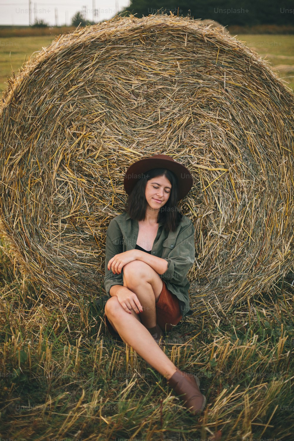 Beautiful stylish woman in hat sitting at hay bale in summer evening field. Happy young female relaxing at haystacks, summer vacation in countryside. Tranquility, countryside slow life
