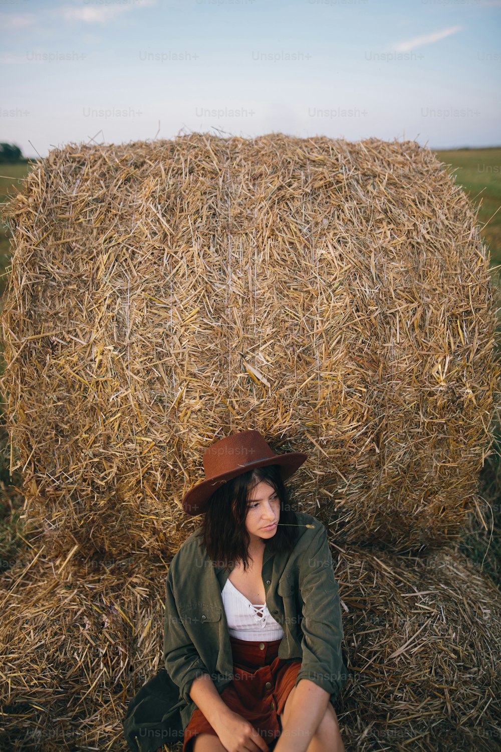 Beautiful stylish woman in hat sitting at hay bale in summer evening field. Happy young female relaxing at haystacks, summer vacation in countryside. Tranquility, countryside slow life