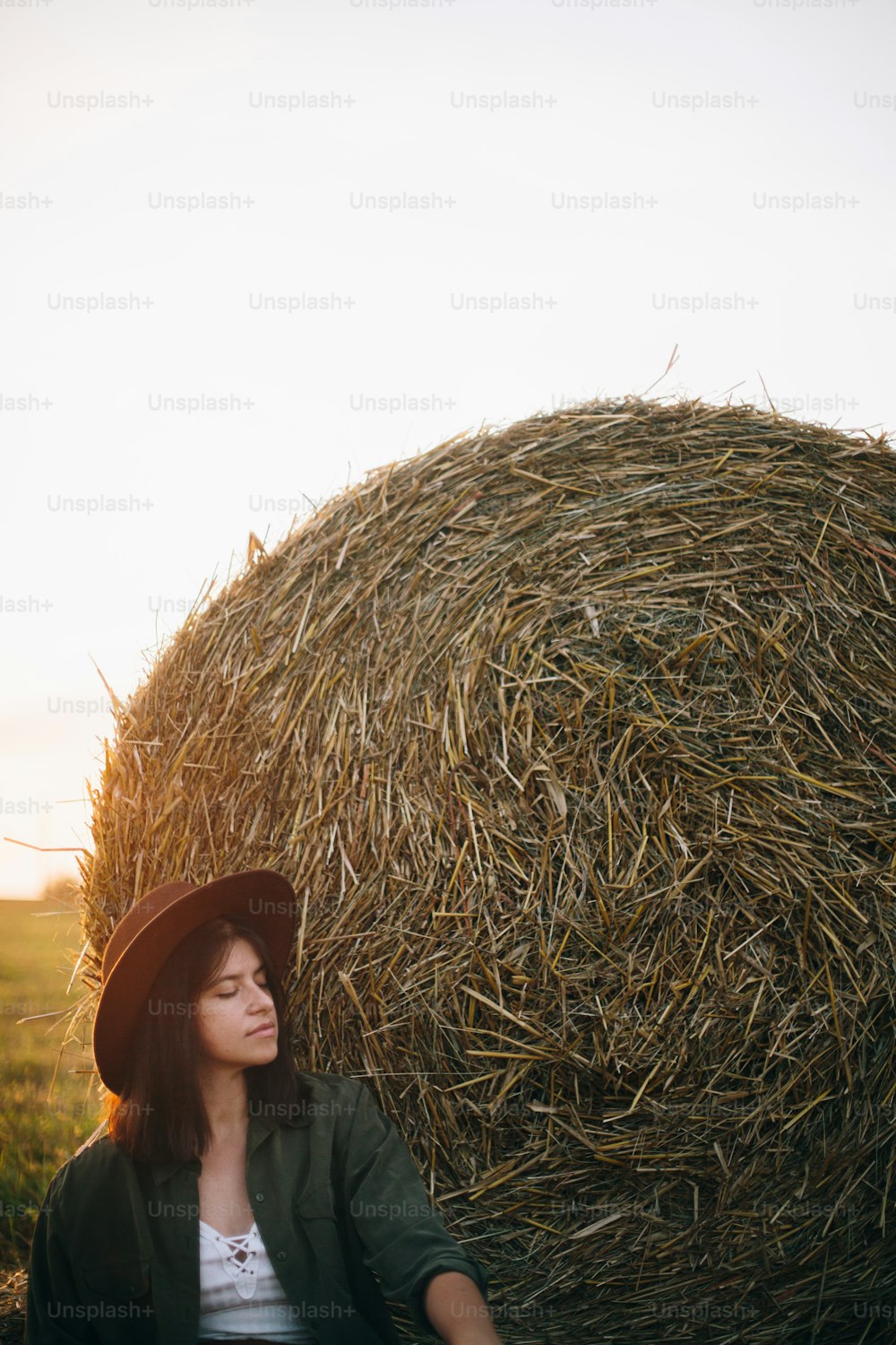 Beautiful stylish woman in hat sitting at haystacks enjoying evening in sunny summer field. Atmospheric tranquil moment in countryside. Young female relaxing at hay bale in warm sunset