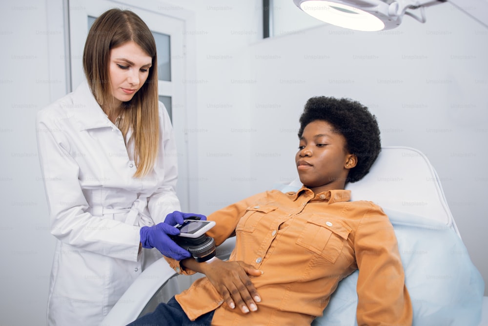 皮膚がんと黒色腫の予防。専門の医師の皮膚科医は、脱毛鏡検査用の最新の装置の助けを借りて、アフリカの女性の腕にある患者のほくろを調べます